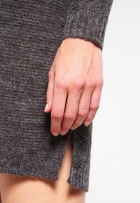 Pull en maille grise avec une finition texturée, doté d'une fente sur le côté. Gros plan sur une main reposant sur le tissu, mettant en valeur la douceur et l'épaisseur.