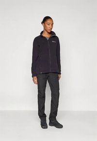 Naine seisab, seljas must lukuga fliistagine, mustad püksid ja mustad matkajalanõud, lihtsa heleda tausta ees.