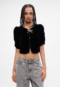 Top corto in velluto nero con maniche a palloncino, dotato di un'apertura frontale legata con un nastro decorativo argentato. Indossato con pantaloni in denim grigi.