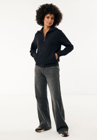 Chaqueta negra con cremallera y cuello alto, hecha de una tela suave. Combinada con jeans sueltos de color gris oscuro y zapatillas negras.