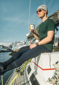 En man som bär en grön t-shirt, marinblå leggings och svarta strumpor sitter på en segelbåt och håller en vit behållare. Snöiga berg i bakgrunden.