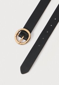 Ceinture en cuir noir avec boucle ronde en or martelé et plusieurs trous d'ajustement sur un fond clair.