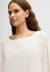 Top di colore crema, texturizzato, con ampia scollatura a barca e maniche ampie e fluttuanti, caratterizzato da una leggera lucentezza e da elementi di design minimalisti.
