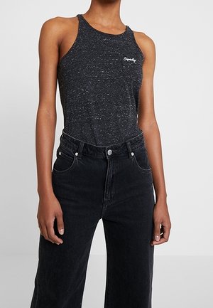Frau trägt ein dunkelgraues ärmelloses Tanktop mit kleinem weißem Logo auf der Brust und hoch taillierte schwarze Jeans.