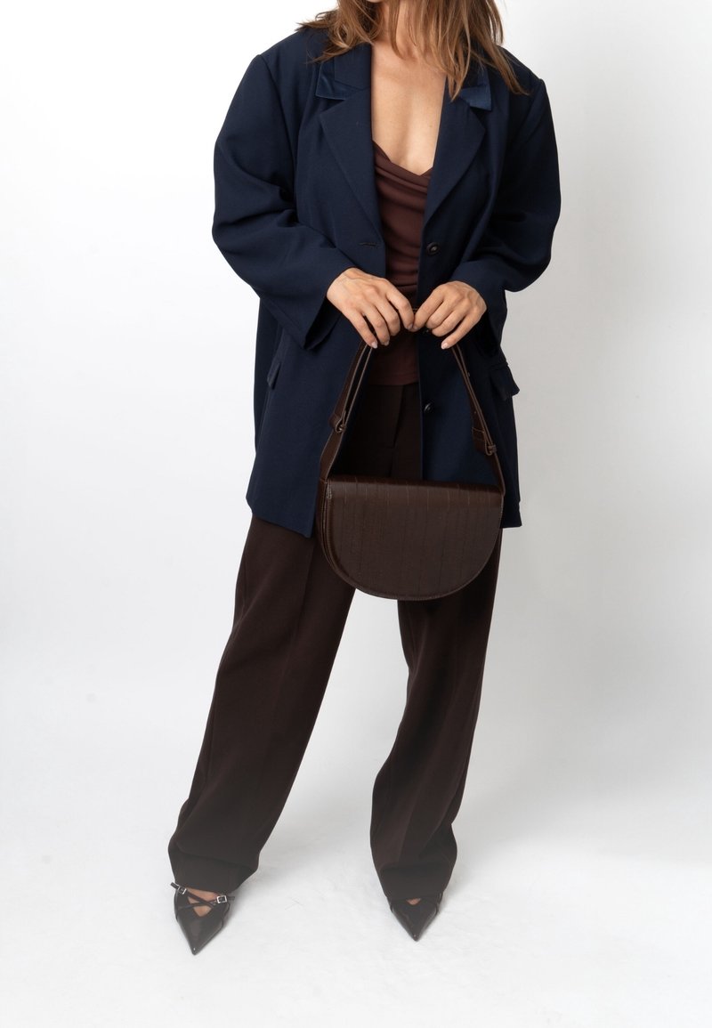Femme portant un blazer bleu marine, un haut marron et un pantalon, tenant un sac à main brun foncé avec des chaussures noires pointues sur fond blanc.