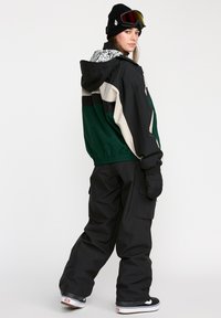 Skijakke i grøn, sort og beige med elastiske manchetter, parret med sorte bukser. Har en mønstret hætte og sneskygge.