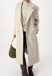 Cappotto doppiopetto in misto lana beige con rever a scendere, tasche laterali e vestibilità rilassata, abbinato a pantaloni marroni scuri e borsa verde.