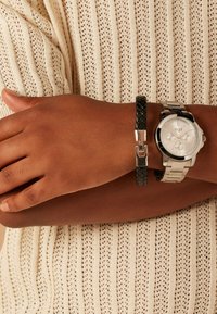 Silver analog watch with a textured white face, accompanied by a black braided leather bracelet. Worn on a hand against a beige knit fabric.