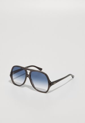 Gafas de sol con montura cuadrada extragrande en plástico marrón oscuro, con lentes azules degradados y acento de logo dorado en el costado.