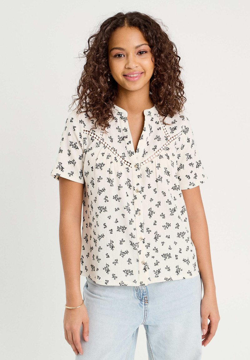 Short-sleeve blouse in cream with black floral print, button front, and lace detailing along the yoke, made from lightweight fabric.