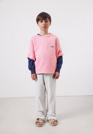 Zēns stāv pret vienkāršu fonu, valkājot rozā īsām piedurknēm džemperi virs tumši zilām garām piedurknēm, gaiši pelēkas bikses un bēšas sandales.