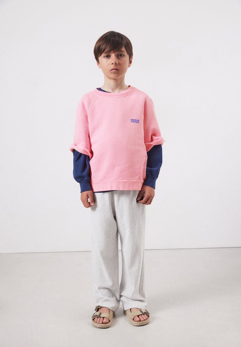 Garçon debout devant un fond uni, portant un sweat-shirt rose à manches courtes sur un sous-pull bleu marine à manches longues, un pantalon gris clair et des sandales beiges.