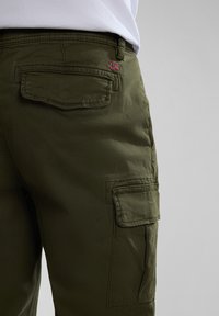 Olivengrüne Cargo-Hosen mit mehreren Taschen, ausgestattet mit einem kleinen bestickten Flaggenmotiv auf der Gesäßtasche, gefertigt aus strukturiertem Stoff.