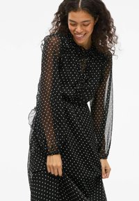 Robe noire à pois avec des manches longues transparentes et un col froncé. Comprend une fermeture à cordon, un tissu fluide et une texture douce.