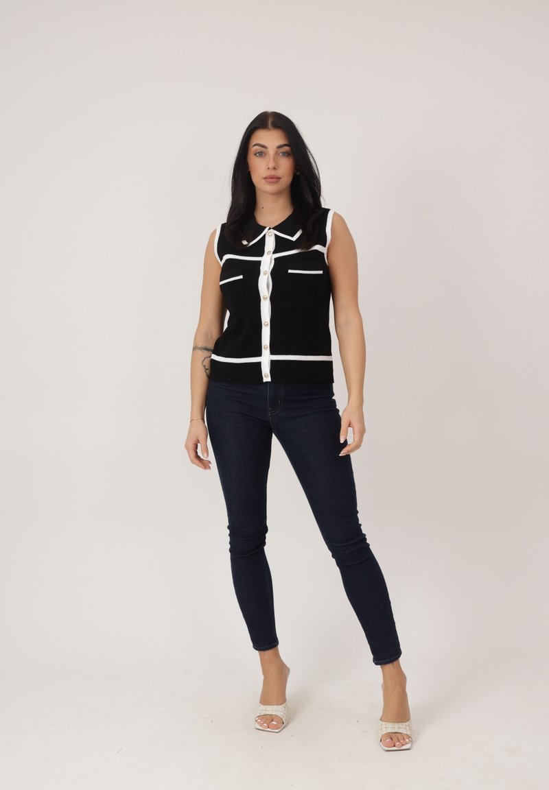 Frau steht vor einem schlichten Hintergrund und trägt ein schwarzes ärmelloses Oberteil mit weißem Rand, dunkle enge Jeans und weiße offene Sandaletten.