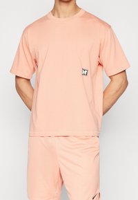 Light pink short-sleeve t-shirt featuring a textured pattern and a small black and white logo patch on the left chest.