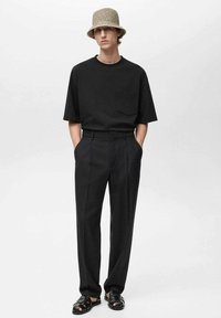 Sort oversized t-shirt med lomme, skræddersyede sorte bukser og sorte sandaler. Modellen bærer en lysfarvet bøttehat med mønster.