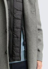 Grauer Außenmantel mit strukturiertem Muster, ausgestattet mit einem schwarzen isolierten Innenfutter, einem Reißverschluss und einem hellblauen Pullover, der darunter sichtbar ist.