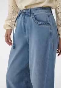 Lyseblå denim bukser med vid ben, bælte i taljen og indsnitdetaljer. Har sidelommer og en glat tekstur.