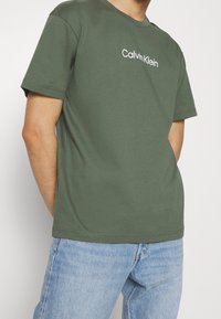 Oliwkowa bawełniana koszulka z okrągłym dekoltem, krótkimi rękawami i białym logo "Calvin Klein" na piersi, zestawiona z jasnoniebieskimi dżinsami.