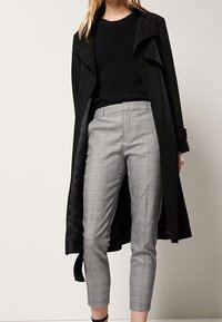 Zwarte trenchcoat over een gestructureerde zwarte trui, gecombineerd met grijs geruite broek die net boven de enkel is geknipt.