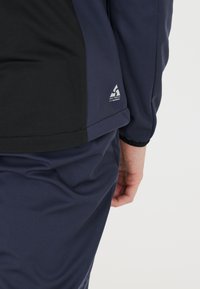 Marinefarbener Sportoberteil mit schwarzen Akzenten, schlanker Schnitt, glatter Textur und kleinem Logo-Patch am unteren Saum.