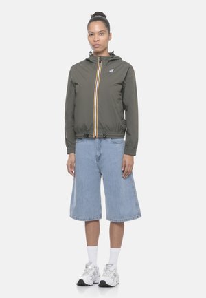 Donna che indossa una giacca con cappuccio verde oliva con zip, pantaloncini denim azzurri a gamba larga, calzini bianchi e scarpe da ginnastica bianche, in piedi su sfondo bianco.