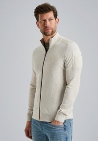 Beige Hoodie mit Reißverschluss, hochgeschlossenem Kragen, gerippten Bündchen und einer aufgesetzten Tasche am Ärmel. Weiche Textur; taillierte Passform.