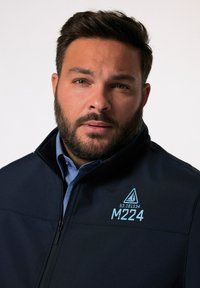 Giacca softshell blu scuro con chiusura a zip, caratterizzata da grafiche stampate in blu, tra cui un triangolo e "M224." Finitura testurizzata visibile.