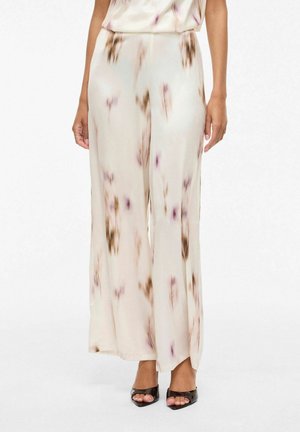 Kvinne iført kremfargede bukser med vide ben og dempede brune og lilla vannfargelignende flekker, kombinert med svarte åpne tå-hæler.