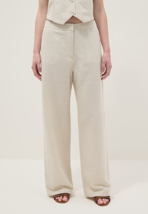 Pantalones beige de pierna ancha combinados con un chaleco corto beige abotonado y sandalias marrones de punta abierta sobre un fondo liso.