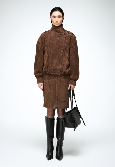 Vrouw draagt een bruine suède jas en bijpassende rok, zwarte laarzen tot aan de knie, houdt een zwarte handtas vast, staat tegen een effen achtergrond.