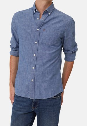 Uomo che indossa una camicia azzurro chiaro con bottoni e maniche lunghe con taschino sul petto e jeans blu scuro, le maniche arrotolate fino ai gomiti.