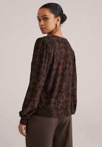 Rjava bluza s teksturiranim cvetličnim vzorcem v črni barvi, z dolgimi rokavi in okroglim izrezom, v kombinaciji z rjavimi hlačami.