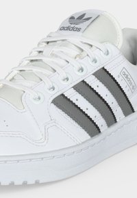 Λευκό αθλητικό παπούτσι Adidas με τρεις μαύρες ρίγες στο πλάι, λευκά κορδόνια και λογότυπο Adidas στη γλώσσα και το πλευρικό πάνελ.