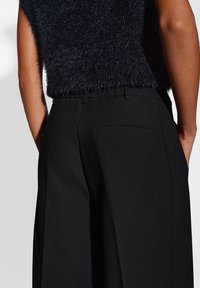 Pantalons larges noirs avec une texture lisse, comportant une poche arrière et une taille élastique, associés à un haut en tricot foncé et duveteux.