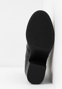 Chaussure en cuir noir avec une semelle en caoutchouc texturée, un talon légèrement surélevé et un dessus lisse avec un subtil détail de couture. Taille marquée à l'intérieur.