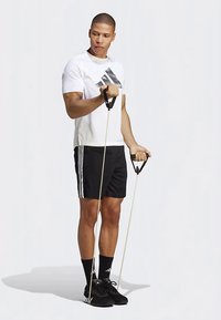 Pantalones cortos deportivos negros con rayas blancas, una camiseta blanca y zapatillas negras. Sosteniendo bandas de resistencia con asas. Fondo neutro.