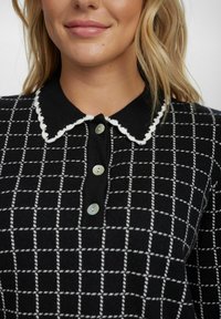 Polo shirt à carreaux noir et blanc avec un col scallopé et trois boutons décoratifs en matériau nacré.