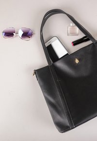 Schwarze Lederhandtasche mit Smartphone darin, daneben eine Sonnenbrille und Make-up-Artikel einschließlich Lippenstift und Parfüm auf einer hellen Oberfläche.