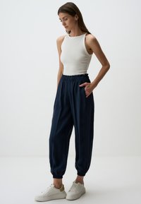Marineblaue, taillierte Hose mit elastischem Bund und seitlichen Taschen, kombiniert mit einem weißen, figurbetonten Tanktop und weißen Sneakers. Glatte Stofftextur.