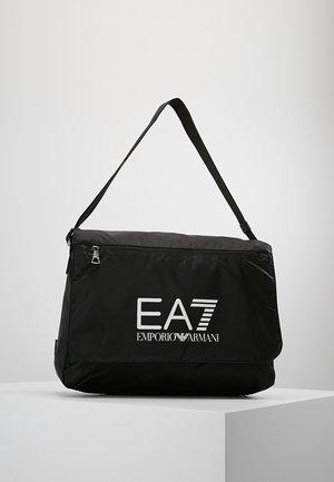 Bolso negro de hombro con correa ajustable, bolsillo delantero con cremallera y logo blanco de EA7 Emporio Armani en la solapa, colocado sobre una superficie blanca.