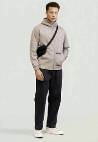Felpa beige con zip e tasca frontale, abbinata a jeans neri a gamba dritta e sneakers bianche. Borsa a tracolla nera con accentuazione del logo.
