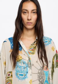 Durchsichtige Hemdbluse aus leichtem beigen Stoff mit bunten Druckmotiven, darunter Gesichter, Formen und florale Akzente.