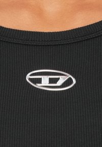 Melna ribota tanks, kurā pie kakla ir sudraba ovāls logotips ar stilizētu "D" centrā.