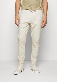 Pantalones de algodón beige con cintura ajustable, corte Slim y bolsillos laterales, combinados con zapatillas altas blancas y una camiseta verde claro.
