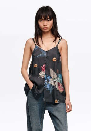Mujer con el cabello negro liso, vestida con una camiseta negra abotonada con patrones coloridos de flores y aves, combinada con jeans azules holgados.