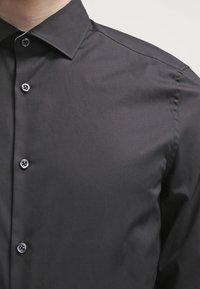 Camicia nera realizzata in tessuto morbido, con design a bottone, colletto a punta e bottoni neri a contrasto.