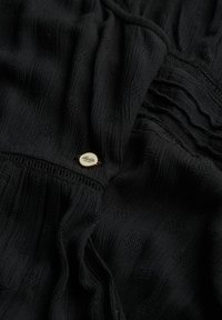 Czarna tkanina z teksturowym wzorem z wypukłymi kropkami. Zdobiona złotym guzikiem z misternym detalem. Miękka i lekko zmarszczona struktura.