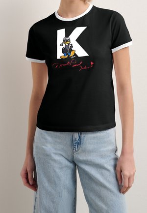 Vrouw draagt een zwart T-shirt met witte kraag en mouwen, met een grote witte "K", Donald Duck-afbeelding en rode tekst eronder. Lichtblauwe jeans ook zichtbaar.
